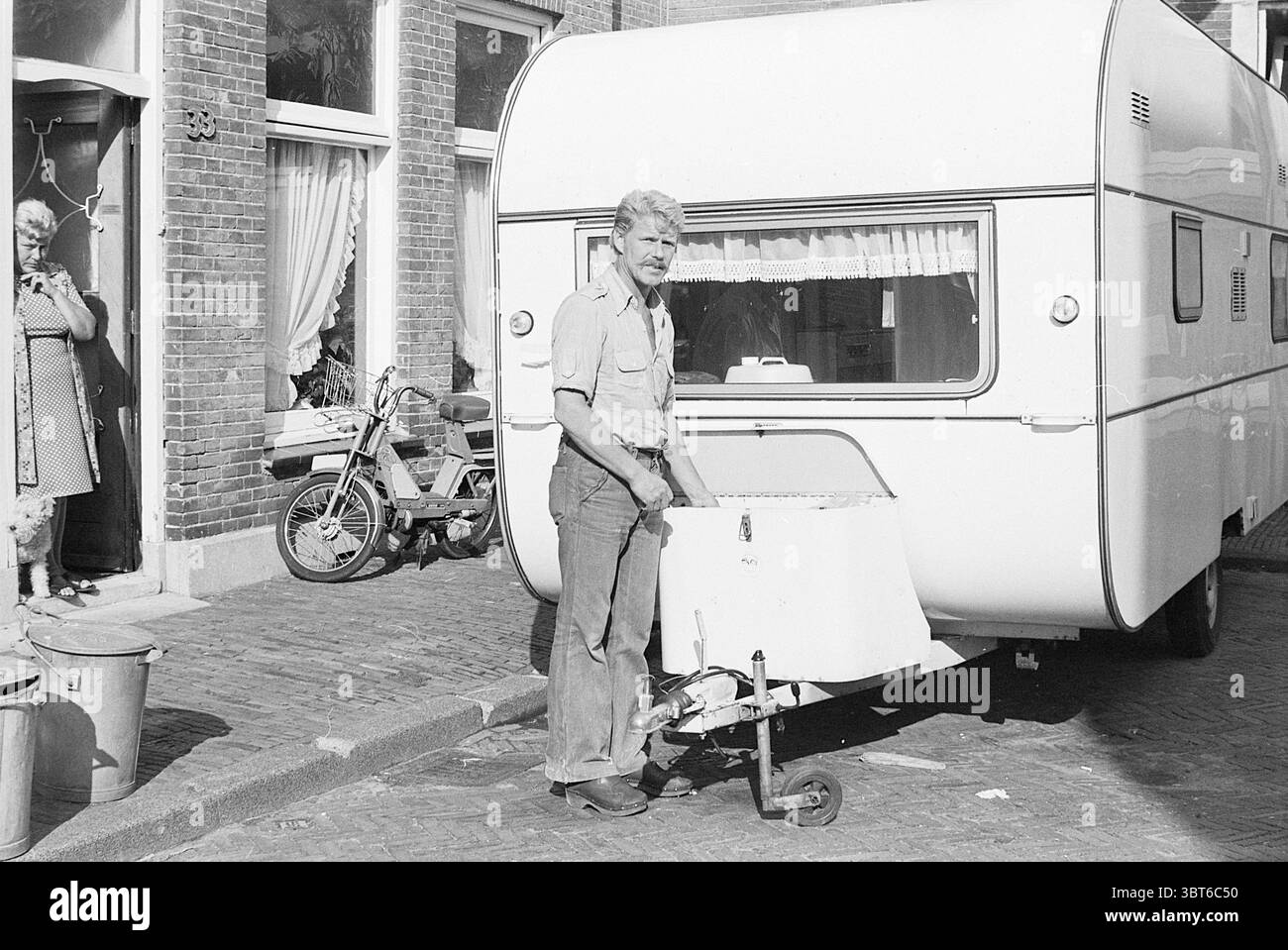 Sig. Goebert presso Caravan Persons, Whizgle News, Dutch Desk, Paesi Bassi, 1950 - 2000 il 03-08-1976. L'immagine contiene questi argomenti. La scena presenta un angolo di strada soleggiato con una carovana bianca e luminosa ben parcheggiata. La roulotte ha un esterno liscio e lucido e un design semplice e moderno con bordi rotondi, che creano un senso di apertura. Di fronte a essa, un uomo di mezza età sta in piedi, afferrando la maniglia di una piccola piattaforma a ruote collegata alla roulotte. Indossa una camicia a maniche corte di colore chiaro e appare concentrato, con un leggero solco sulla fronte. Dietro di lui, l'ambientazione è caratteristica Foto Stock