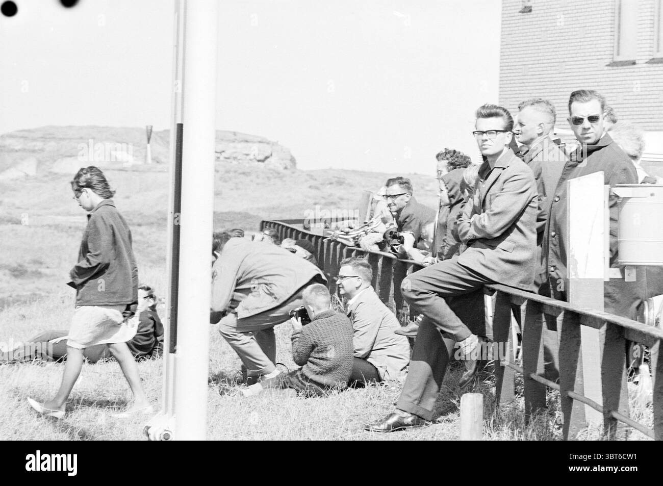 I rimorchiatori di Wijsmuller portano un carro di perforazione a Wijsmuller, Whizgle News, Dutch Desk, Paesi Bassi, 1950 - 2000 su 26-08-1963. L'immagine contiene questi argomenti. La scena cattura un raduno di persone su terreni erbosi, con un mix di abbigliamento casual e più formale. In primo piano, un uomo con gli occhiali è seduto casualmente su una ringhiera, trasudando un'aria di rilassata sicurezza. Il suo abbigliamento è composto da un abito scuro che contrasta con la luce circostante. Nelle vicinanze, molti altri sono impegnati in varie attività, alcune accovacciate e focalizzate su un punto di interesse specifico. La folla è composta da uomini Foto Stock