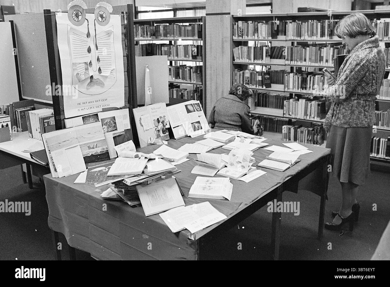 Destroyed book Exhibition Beverwijk Library Books and booksellers, Whizgle News, Dutch Desk, Paesi Bassi, 1950 - 2000 il 07-02-1977. L'immagine mostra questi argomenti. In una spaziosa biblioteca, la scena presenta un tavolo centrale ricoperto di vari tipi di carta, libri e materiali stampati, appoggiato su un panno rosso. Gli elementi sembrano essere un mix di opuscoli, volantini e documenti, possibilmente correlati a un evento informativo o espositivo. A sinistra, una persona è seduta, impegnata a guardare attraverso i materiali sul tavolo, la schiena si rivolge allo spettatore. Indossano un outfit classico, aggiungendo Foto Stock
