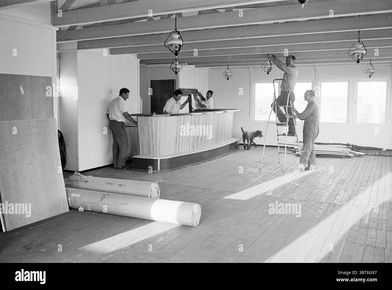 Costruzione di una mensa., Whizgle News, Dutch Desk, Paesi Bassi, 1950 - 2000. Questi sono gli argomenti nell'immagine. La scena raffigura un interno spazioso in fase di ristrutturazione, caratterizzato da un senso di attività e trasformazione. La sala presenta travi in legno sul soffitto, che donano un fascino rustico con una luce naturale. In primo piano, diversi rotoli di materiale, possibilmente tappeto o linoleum, giacciono casualmente sul pavimento, suggerendo il lavoro in corso. Verso il centro, un banco ricevimento in legno, parzialmente oscurato dalla vista, funge da punto focale. Dietro, ci sono due individui Foto Stock