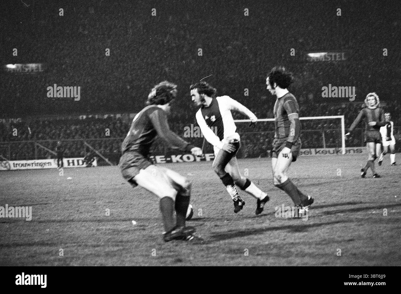 Partita di calcio Ajax - Bayern Monaco Amsterdam Nederland., Whizgle News, Dutch Desk, Paesi Bassi, 1950 - 2000. L'immagine include questi argomenti. La scena cattura un momento intenso di una partita di calcio sotto un cielo scarsamente illuminato, mettendo in risalto l'atmosfera elettrica dello stadio. In primo piano, due giocatori sono impegnati in un gioco attivo. Un giocatore, indossando un'uniforme scura, accovacciato leggermente con un'espressione mirata, pronto a riprendere possesso della palla. L'altro, indossando una maglia bianca con finiture scure, passa oltre, evitando abilmente un tackle. I suoi capelli vengono leggermente arruolati durante le manovre Foto Stock