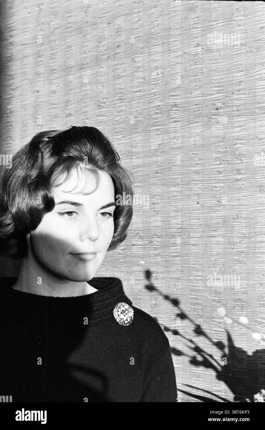 Signora Broeren arrangiatore di fiori giapponese Persons, Whizgle News, Dutch Desk, Paesi Bassi, 1950 - 2000 il 21-01-1963. Questi argomenti vengono visualizzati nell'immagine. In un ambiente con luce soffusa, una giovane donna si erge su uno sfondo testurizzato che ha un aspetto in fibra naturale, a trasmettere un'atmosfera calda e rustica. I toni tenui dello sfondo contrastano con i suoi abiti scuri, che hanno una leggera lucentezza, esaltando la raffinatezza del suo look. La sua acconciatura è chic e curata, per aggiungere un fascino vintage alla scena. Una spilla delicata, forse in metallo o vetro, è fissata al suo colletto, gatto Foto Stock