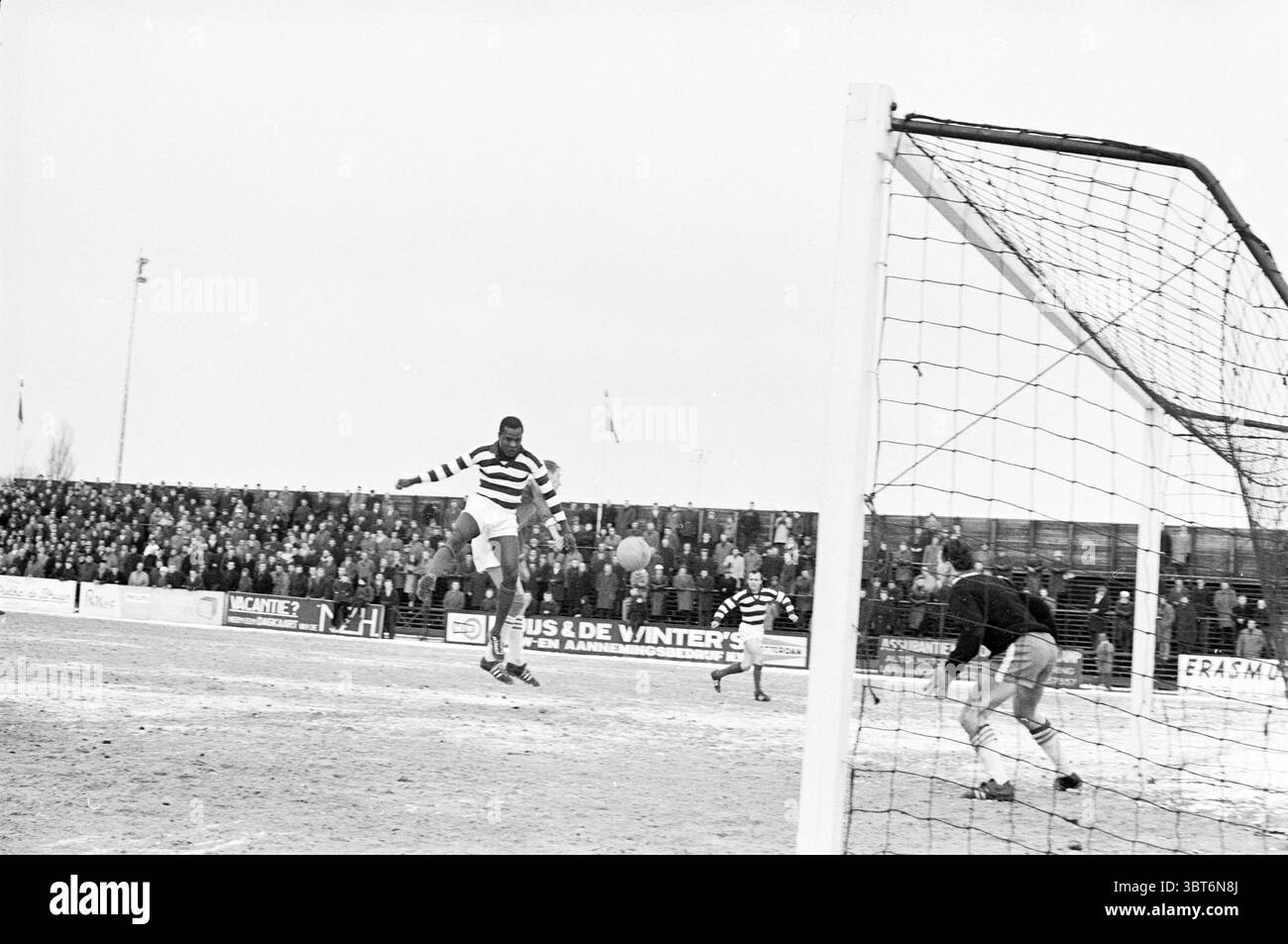 R.C.H. - Blue and White Football, Whizgle News, Dutch Desk, Paesi Bassi, 1950 - 2000 il 03-02-1963. Questi argomenti sono mostrati nell'immagine. In una scena sportiva dinamica, un giocatore con una maglia a strisce salta in aria, sapientemente pronto a fare un gioco decisivo sul pallone da calcio. Il giocatore mette in mostra un mix di agilità e determinazione, con una forte concentrazione sul viso. Le strisce distintive della maglia contrastano con le sfumature del cielo nuvoloso, creando un effetto visivo sorprendente. Intorno al giocatore, l'obiettivo è ben visibile, con i suoi pali bianchi che incorniciano l'azione. La compensazione Foto Stock