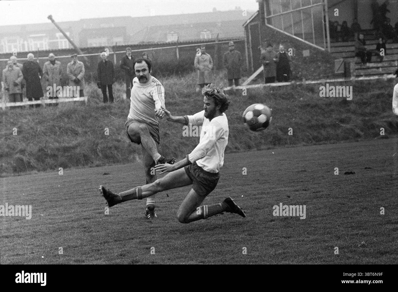 Zandvoortmeeuwen - SV Ilpendam Football Zandvoortse Meeuwen Halfweg, Whizgle News, Dutch Desk, Paesi Bassi, 1950 - 2000 il 30-11-1975. Questi argomenti sono mostrati nell'immagine. La scena cattura un momento energetico su un campo di calcio erboso dove due giocatori sono attivamente impegnati in una partita competitiva. Un giocatore, vestito con una maglia di colore chiaro, tenta abilmente di calciare la palla mentre gli si avvicina. La sua espressione trasmette determinazione, sopracciglia a fuoco. Il giocatore avversario, che indossa una maglia più scura, è in movimento, una gamba sollevata mentre tenta di bloccare o intercettare la palla. Suo Foto Stock