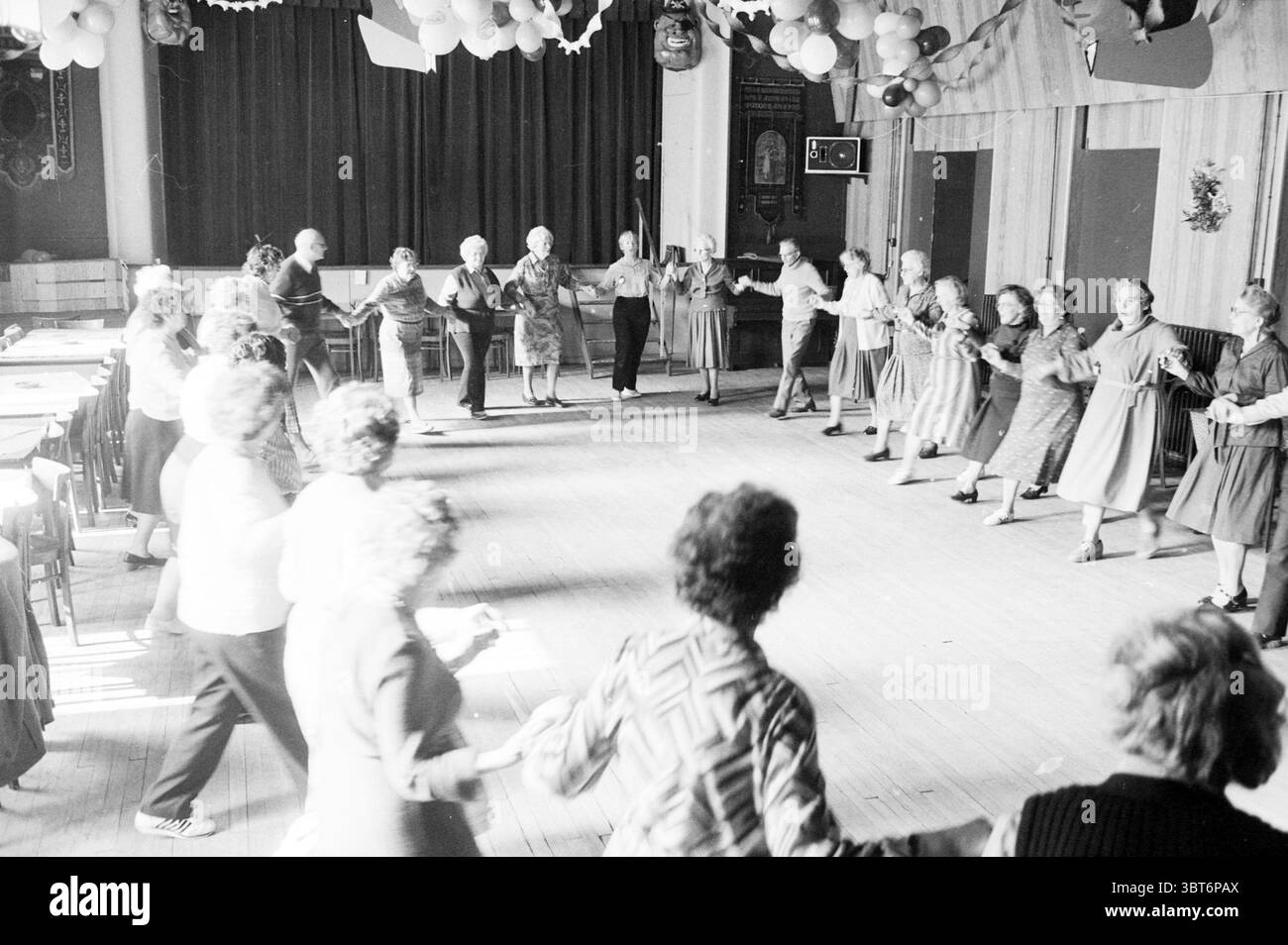 Danza popolare per gli anziani Lisse danza popolare Lisse, Whizgle News, Dutch Desk, Paesi Bassi, 1950 - 2000 il 12-03-1984. Questi argomenti sono mostrati nell'immagine. In un ambiente interno vivace, la scena si snoda con un gruppo di persone impegnate nella danza, creando un'atmosfera piena di gioia ed energia. Lo spazio presenta un grande pavimento in legno, lucidato e riflettente, che fornisce un'area invitante per il movimento. Le pareti sono adornate da tendaggi e palloncini colorati sono appesi al soffitto, aggiungendo un tocco di festa. Intorno ai bordi della scena, le sedie sono allineate in modo ordinato, suggerendo questo Foto Stock