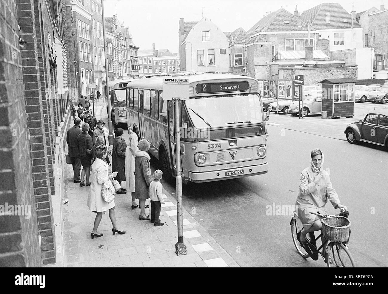 Fermata dell'autobus Damstraat Noord Zuid Hollandse Vervoersmaatschappij N.V. Haarlem Damstraat Paesi Bassi, Whizgle News, Dutch Desk, Paesi Bassi, 1950 - 2000 on 31-10-1969. L'immagine mostra questi argomenti. La scena si snoda in un ambiente urbano caratterizzato da un mix di architettura antica e di metà secolo. Su un lato della strada, sono parcheggiati due grandi autobus d'epoca, i loro colori vivaci oscurati da un filtro monocromatico, creando un'aria di nostalgia. L'autobus più vicino allo spettatore mostra in modo prominente il numero "2" e una destinazione del percorso che indica il transito locale, suggerendo che si tratta di un mezzo occupato Foto Stock