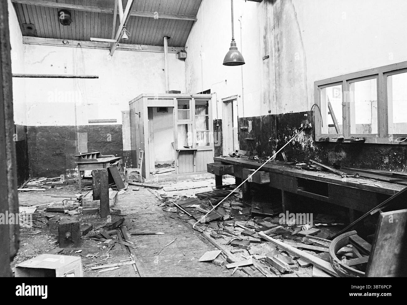 Demolizione ecc. garage Voorburg Noord Zuid Hollandse Vervoersmaatschappij N.V. NZ, Whizgle News, Dutch Desk, Paesi Bassi, 1950 - 2000 il 23-03-1965. L'immagine include questi argomenti. La scena presenta una spaziosa officina abbandonata caratterizzata da un mix di elementi industriali e rustici. Il soffitto alto è decorato con travi a vista, che aggiungono un tocco di atmosfera grezza. Una luce soffusa a sospensione pende dal soffitto, proiettando ombre morbide in tutta la stanza. Il pavimento è disseminato di detriti, tra cui legno scheggiato e frammenti di metallo, a indicare un lungo periodo di abbandono. Al centro, una grande banchina da lavoro usurata Foto Stock