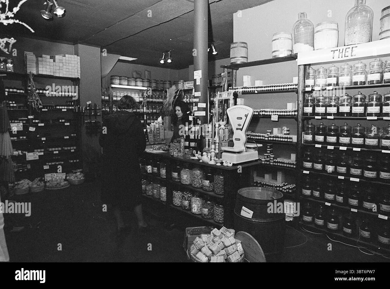 Negozio di erbe Barrevoetestraat negozi e chioschi Haarlem Barrevoetestraat Paesi Bassi, Whizgle News, Dutch Desk, Paesi Bassi, 1950 - 2000 il 07-01-1977. Questi sono gli elementi dell'immagine. La scena si snoda in un pittoresco negozio vecchio stile che trasuda un fascino nostalgico. L'interno è pieno di ripiani rivestiti con vari vasetti e contenitori, creando un ricco arazzo di forme e dimensioni. Molti di questi vasi di vetro sono pieni di un assortimento di articoli colorati, che vanno da erbe e spezie vibranti a frutta secca e dolci, il tutto meticolosamente organizzato. Le pareti sono di un colore morbido e tenue Foto Stock