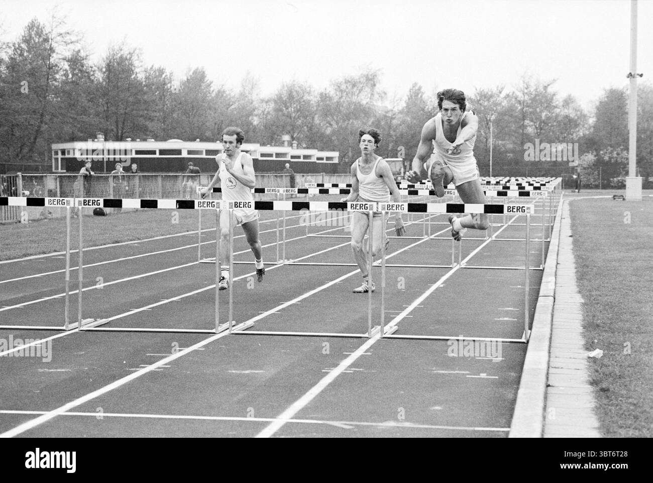 Gara di hurdle racing., Whizgle News, Dutch Desk, Paesi Bassi, 1950 - 2000. L'immagine include questi argomenti. La scena raffigura un gruppo di tre atleti impegnati in una gara di ostacoli su una pista. Sono posizionati in una posizione dinamica e competitiva mentre attraversano gli ostacoli. Gli atleti sono dotati di uniformi che si adattano perfettamente alla forma, che ne esaltano l'aspetto snello durante lo sprint in avanti. La pista è caratterizzata da linee chiare e presenta più ostacoli, che sono distanziati uniformemente lungo le corsie. La superficie è scura, probabilmente asfalto, in contrasto con i colori più chiari dell'AT Foto Stock