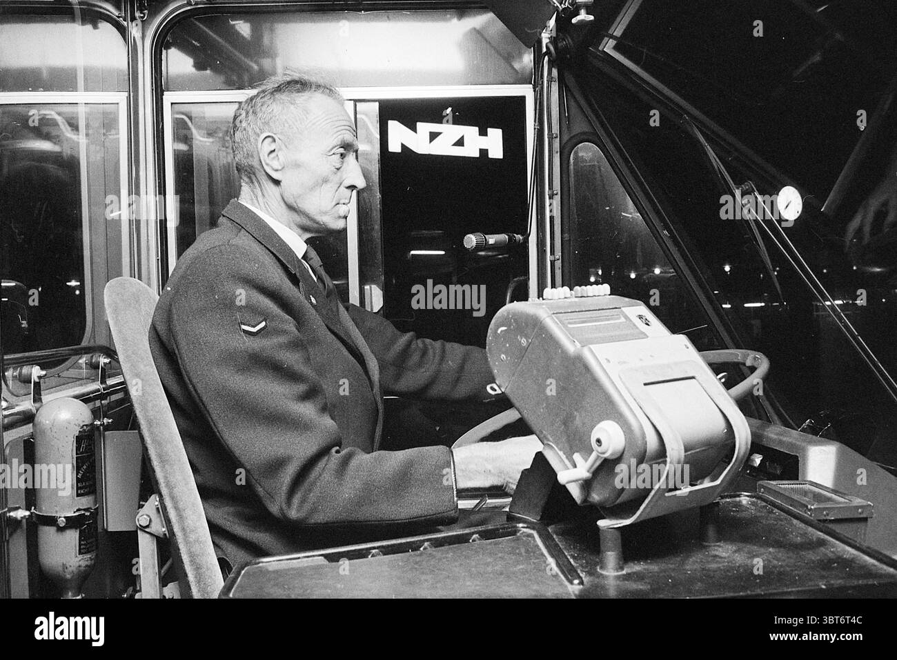 Autista al volante Noord Zuid Hollandse Vervoersmaatschappij N.V. NZ, Whizgle News, Dutch Desk, Paesi Bassi, 1950 - 2000 il 15-10-1970. Questi argomenti vengono visualizzati nell'immagine. La scena vede un uomo maturo seduto ai comandi di un veicolo, che trasuda un'aria di concentrazione e professionalità. Ha un'espressione severa ma concentrata, che suggerisce un profondo senso di responsabilità per il suo compito. Il suo abbigliamento è formale, suggerendo che detiene una posizione di autorità o esperienza nel suo ruolo. Gli interni del veicolo sono strutturati e funzionali e presentano una serie di strumenti e quadri di comando Foto Stock