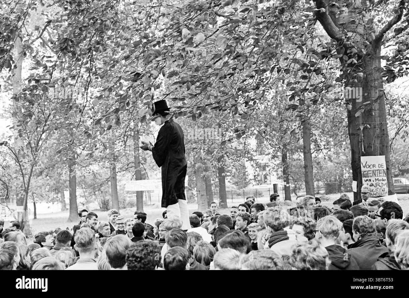 Elezioni del Consiglio scolastico Mendelcollege e Karel van Manderlyceum Giochi giovanili e giovanili scuole, Whizgle News, Dutch Desk, Paesi Bassi, 1950 - 2000 il 11-10-1963. L'immagine contiene questi argomenti. La scena è piena di una folta folla riunita in un parco, circondato da grandi alberi frondosi che creano un baldacchino naturale in alto. L'atmosfera è carica di energia mentre un altoparlante si trova su una piattaforma rialzata, catturando il pubblico con un'espressione animata. Vestito con un lungo cappotto nero e un cappello alto, l'altoparlante trasuda un senso di carisma e autorevolezza. Tra la folla, un mix di individua Foto Stock