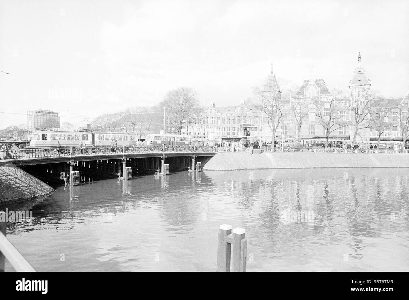 Il fronte del Porto aperto si vede verso la stazione centrale Amsterdam Stationsplein Nederland, Whizgle News, Dutch Desk, Paesi Bassi, 1950 - 2000 il 11-03-1971. Questi argomenti sono mostrati nell'immagine. La scena cattura un tranquillo paesaggio urbano caratterizzato da un ampio corso d'acqua che riflette un mix di luci e ombre. Su un lato dell'acqua si estende un ponte ampio e robusto, costruito con materiali scuri e robusti con pilastri di supporto immersi nell'acqua. Un tram può essere visto muoversi lungo il ponte, trasmettendo un senso di movimento in un ambiente tranquillo. Sullo sfondo, una fila di elega Foto Stock