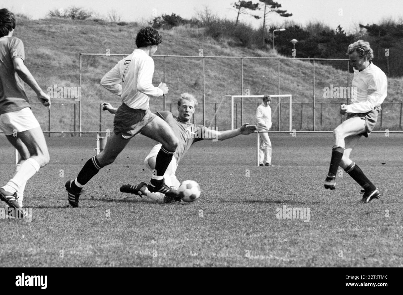 Stormvogels - De Spartaan Football V.S.V. Velsen Stormvogels, Whizgle News, Dutch Desk, Paesi Bassi, 1950 - 2000 il 11-04-1982. L'immagine mostra questi argomenti. La scena cattura un momento vivace su un campo di calcio in erba, dove è in corso una partita animata. In primo piano sono presenti tre giocatori attivamente impegnati nel gioco: Un giocatore con una maglia grigia sta scivolando per terra, tentando di intercettare la palla. La sua espressione è focalizzata, a indicare la sua determinazione nella partita. Alla sua sinistra, un altro giocatore con una maglia blu sembra manovrare abilmente intorno a lui, con la palla vicina t Foto Stock