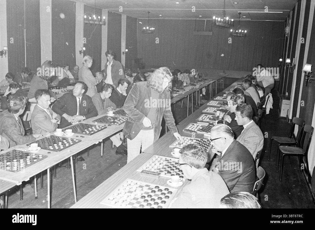 Sijbrandt Checkers simultaneously Checkers, Whizgle News, Dutch Desk, Paesi Bassi, 1950 - 2000 il 21-09-1970. L'immagine include questi argomenti. In una sala spaziosa e ben illuminata, un grande raduno di persone è impegnato nel gioco delle dama. Tavoli lunghi e rettangolari si estendono lungo la stanza, ognuno meticolosamente disposto con scacchiere allestite per il gioco. Giocatori di età variabile siedono su entrambi i lati delle tavole, in profondità, con alcuni inclinati in avanti, mentre altri adottano una postura più rilassata, sorseggiando da piccole tazze. L'arredamento della camera è caratterizzato da pannelli di legno scuro, che creano un caldo voi Foto Stock