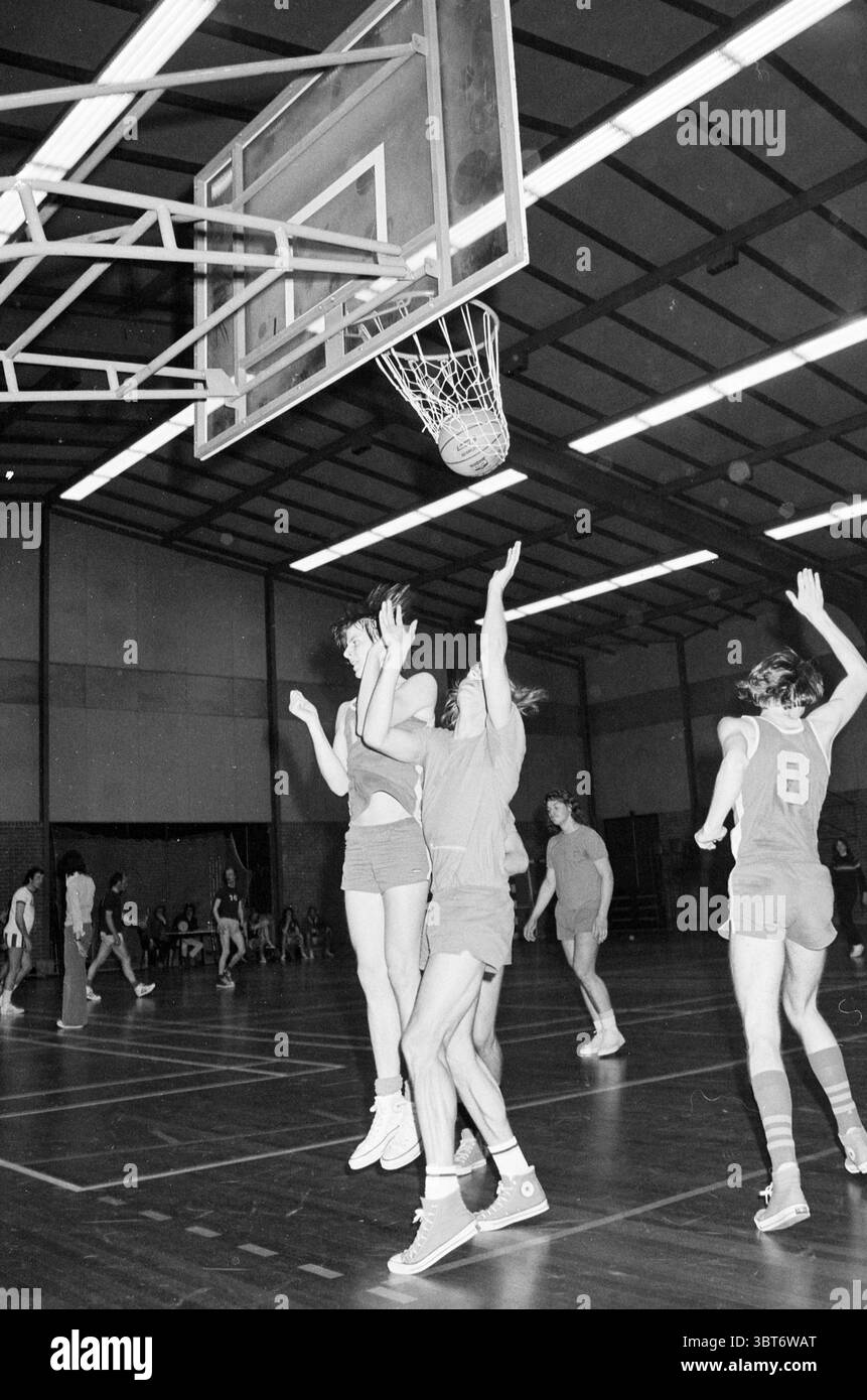Partita di basket., Whizgle News, Dutch Desk, Paesi Bassi, 1950 - 2000. L'immagine include questi argomenti. In una spaziosa palestra interna, l'atmosfera è elettrica con attività. Il pavimento in legno duro riflette una calda luce soffusa dalle luci superiori, creando vivaci luci e ombre. L'ambiente è caratterizzato da un alto soffitto sostenuto da travi a vista che danno un senso di apertura. Un gruppo di atleti è impegnato in una partita di basket vivace, i loro movimenti dinamici e pieni di energia. Giocatori in uniformi vivaci, prevalentemente in tonalità di rosso e blu, saltano e si allungano verso una towerin Foto Stock