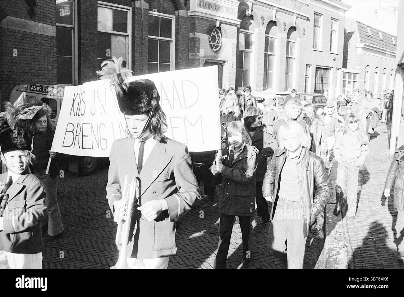 Parata dei bambini Burgwal Haarlem Parades Haarlem Burgwal Paesi Bassi, Whizgle News, Dutch Desk, Paesi Bassi, 1950 - 2000 il 26-10-1974. Questi argomenti vengono visualizzati nell'immagine. In un ambiente urbano vivace, si vede un grande gruppo di bambini che marciano lungo una strada acciottolata. L'atmosfera è piena di determinazione e di scopo. I bambini sono vestiti con vari abiti, tra cui giacche e cappelli, con alcune vivaci piume sportive nel copricapo, che aggiungono un tocco giocoso alla scena. Un ragazzo davanti conduce la processione, tenendo in mano una tromba, vestita con un'uniforme che featu Foto Stock