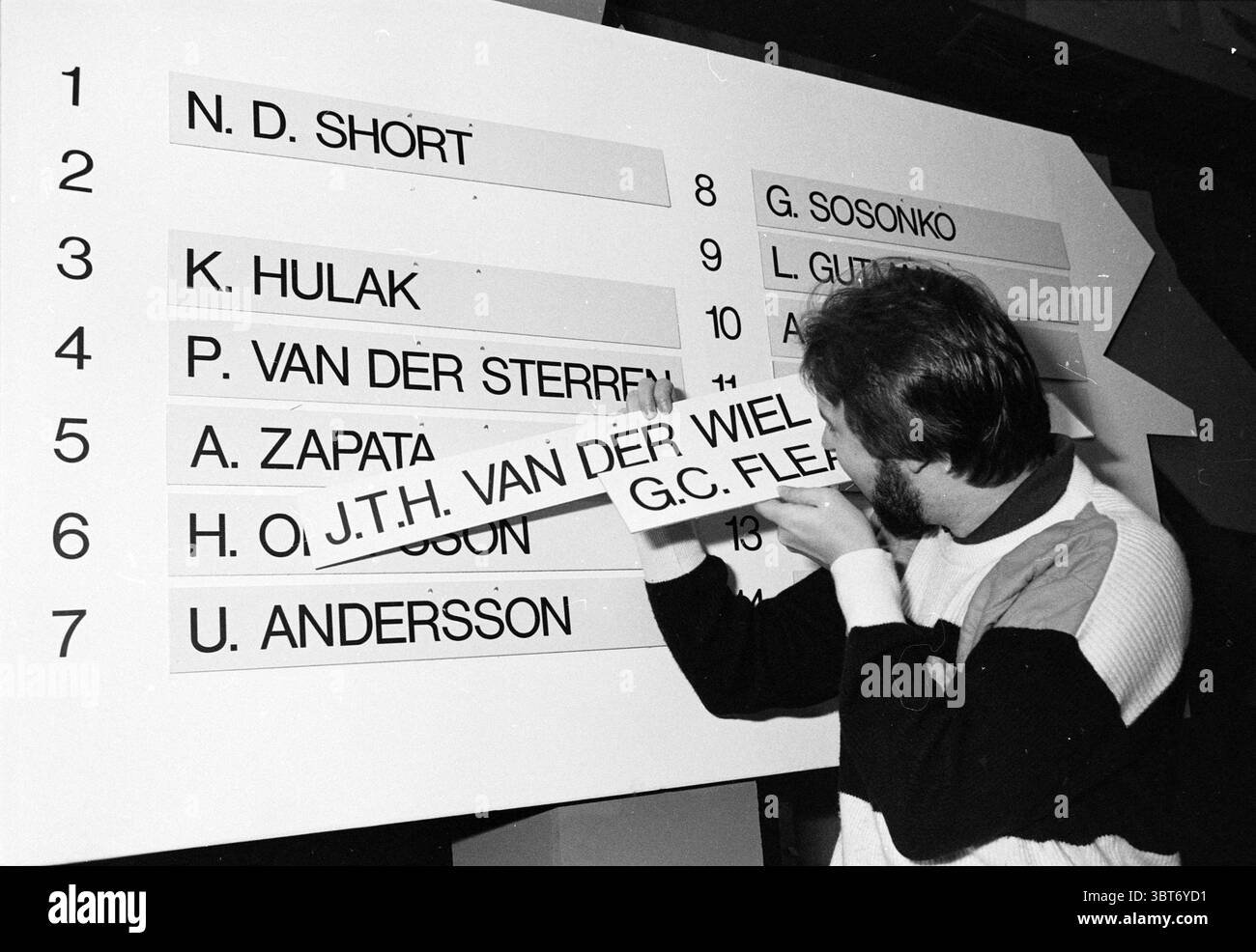 Draw HST - Van de Wiel Blast Furnace Chess Tournament, Whizgle News, Dutch Desk, Paesi Bassi, 1950 - 2000 il 15-01-1987. L'immagine mostra questi argomenti. In un ambiente dinamico, un uomo è impegnato nella regolazione di una grande tavola orientata verticalmente. Lo sfondo è composto da pannelli bianchi con una serie di nomi disposti in un formato elenco. L'individuo, concentrato sul suo lavoro, è vestito con un outfit a contrasto dai colori audaci, con una parte prevalentemente nera completata da accenti luminosi. La sua espressione trasmette concentrazione mentre allinea con cura un pezzo rettangolare, che sembra avere Foto Stock