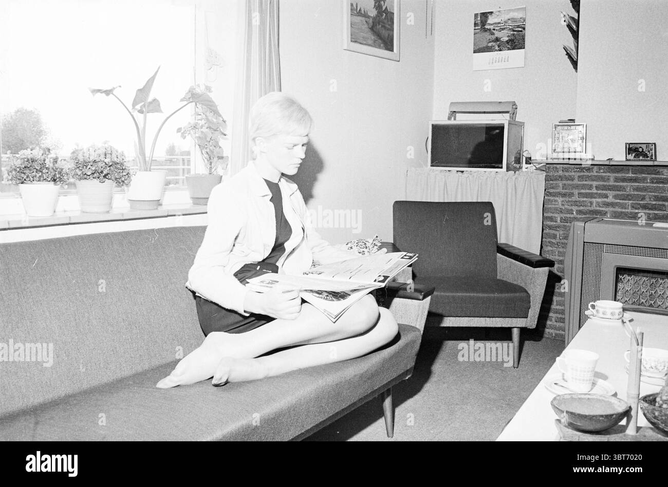 Giovane signora sul divano in soggiorno., Whizgle News, Dutch Desk, Paesi Bassi, 1950 - 2000. Questi argomenti vengono visualizzati nell'immagine. La scena cattura un ambiente interno tranquillo e domestico. Una donna si siede comodamente su un elegante divano scuro, la sua postura rilassata mentre si occupa di diverse riviste e cataloghi in grembo. Ha capelli corti e di colore chiaro semplicemente, che riecheggiano un'estetica minimalista che completa l'atmosfera generale della camera. Il divano stesso e' un tono tenue, possibilmente grigio, che aggiunge un'atmosfera intima ma sofisticata. La camera presenta una varietà di arredi, tra cui una poltrona grigia Foto Stock