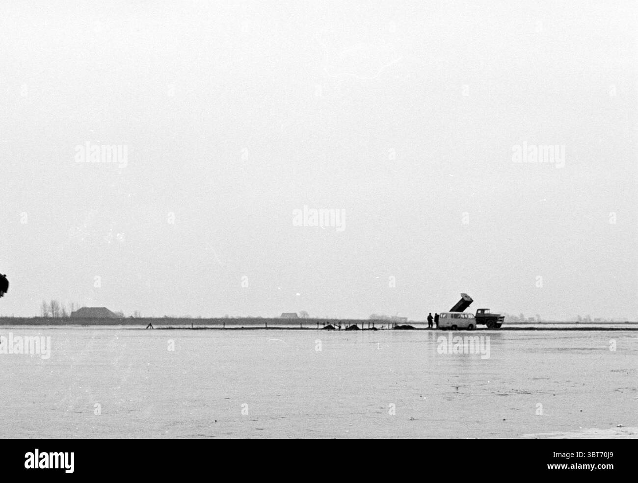 Pattinaggio su ghiaccio naturale, Whizgle News, Dutch Desk, Paesi Bassi, 1950 - 2000. L'immagine mostra questi argomenti. La scena raffigura un vasto paesaggio aperto, dominato da un cielo grigio tenue che suggerisce condizioni meteorologiche sovraffollate. L'orizzonte è basso, con una linea distante di sagome scure, possibilmente casali o alberi, che creano un senso di profondità. In primo piano, un veicolo, probabilmente un vecchio camion, è posizionato vicino al bordo di un corpo d'acqua che riflette i toni cupi del cielo. La superficie sembra calma, con solo sottili increspature che disturbano l'aspetto simile al vetro. Il pianale di carico del carrello è esteso, r Foto Stock