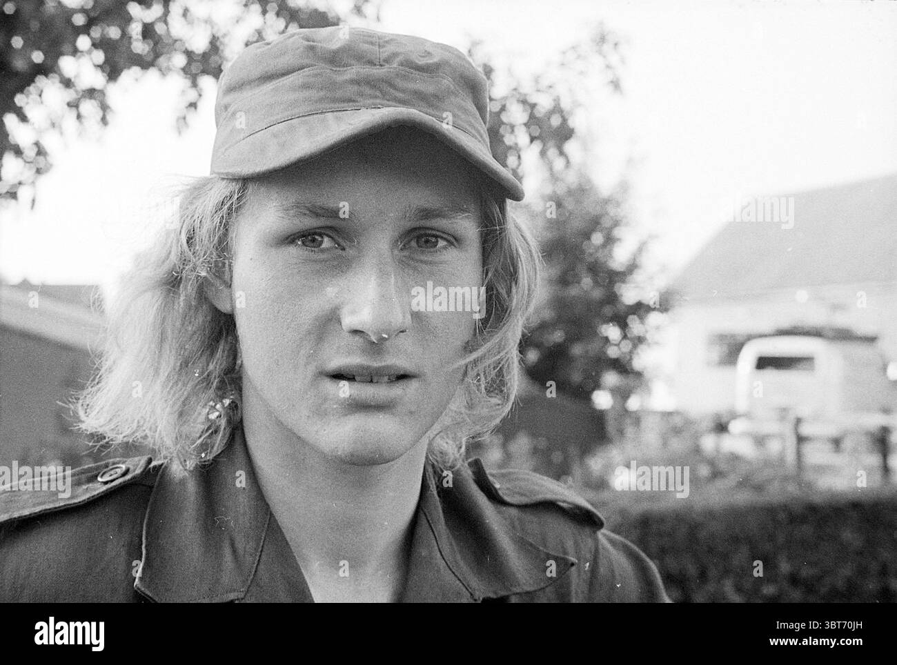 Sig. Waalwijk Persons, Whizgle News, Dutch Desk, Paesi Bassi, 1950 - 2000 il 01-09-1972. L'immagine mostra questi argomenti. La scena presenta un giovane con i capelli biondi leggermente ondulati e con un cappellino in stile militare. La sua espressione è seria, con occhi perforanti che trasmettono un senso di contemplazione o preoccupazione. Lo sfondo è leggermente sfocato, suggerendo un ambiente esterno, possibilmente un'area rurale, con accenni di verde e edifici silenziosi che forniscono un senso di contesto senza travolgere il soggetto. L'illuminazione è naturale e diffusa, probabilmente indica un cielo coperto Foto Stock