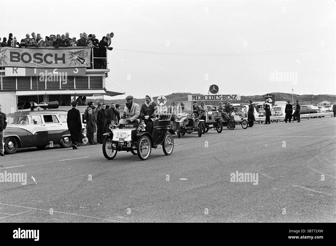 Dimostrazione e corse di auto d'epoca sul circuito di Zandvoort, Whizgle News, Dutch Desk, Paesi Bassi, 1950 - 2000 nel 1962. Questi sono gli elementi dell'immagine. In una scena storica ambientata su un circuito, una linea di veicoli d'epoca occupa il primo piano, ognuno nuvolato in un senso di nostalgia. Le auto sono caratterizzate da un design unico; alcune hanno corpi allungati, mentre altre mostrano una silhouette più compatta. I veicoli, prevalentemente in tonalità silenziose di nero, bianco e grigio, suggeriscono un'epoca di guida passata. Al centro, un'auto si distingue con un conducente che sembra essere in p Foto Stock