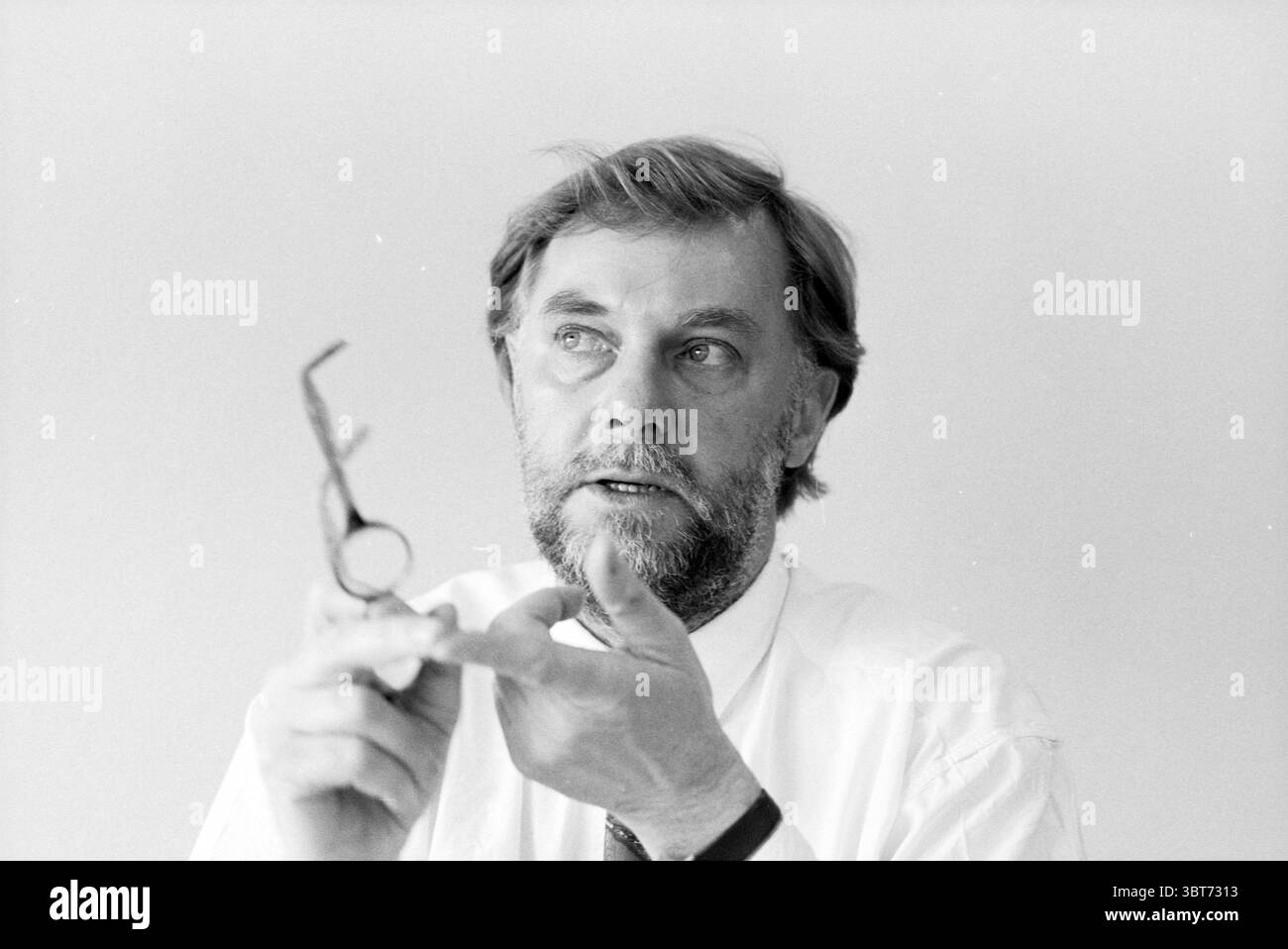 Ken Wilkie Holland Herald Amstelveen Amstelveen, Whizgle News, Dutch Desk, Paesi Bassi, 1950 - 2000 on 28-03-1991. L'immagine contiene questi argomenti. Un uomo di mezza età con un'espressione premurosa occupa il primo piano. Ha una barba ben curata, prevalentemente grigia, che completa i suoi capelli leggermente ruvidi e marrone chiaro. I suoi occhi trasmettono un senso di serietà quando sembra impegnarsi in una discussione o spiegazione. Tiene un paio di occhiali in una mano, la cornice angolare e sottile, in contrasto con la morbidezza delle sue caratteristiche. La tavolozza dei colori è silenziata e presenta principalmente sfumature Foto Stock