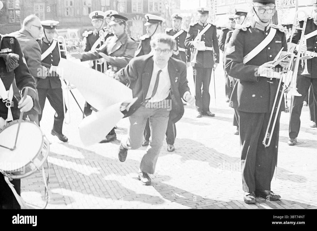 Sommosse alla Marine Corps Chapel Riots, Whizgle News, Dutch Desk, Paesi Bassi, 1950 - 2000 il 18-05-1967. L'immagine contiene questi argomenti. In una vivace scena all'aperto, un gruppo di individui in uniforme occupa il primo piano, vestiti con uniformi nere in stile militare adornate da fasce bianche e spalline lucenti. Alcuni contengono strumenti di ottone, mentre altri suonano percussioni, contribuendo a creare un'atmosfera vivace. La luce del sole bagna la scena, proiettando ombre distinte che migliorano il senso di movimento e di urgenza. Al centro, un uomo con una tuta con un collare aperto tratteggia attraverso la scena, un rotolo Foto Stock