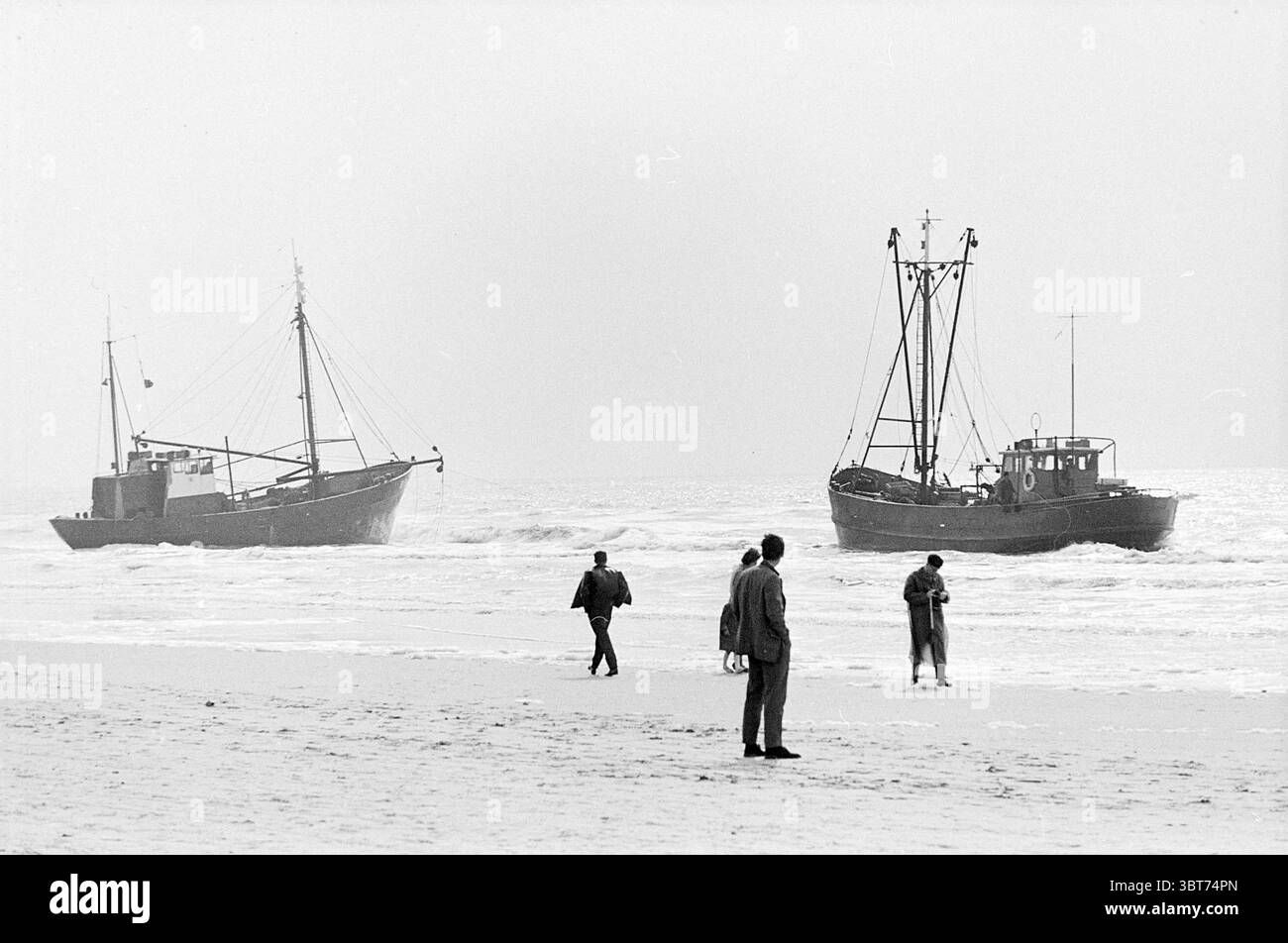 Pescatori di gamberi bloccati sulla spiaggia di Zandvoort BR 22 e TH 32 Beach and Beaches Ships, Whizgle News, Dutch Desk, Paesi Bassi, 1950 - 2000 il 20-05-1963. Questi argomenti vengono visualizzati nell'immagine. La scena raffigura un ambiente costiero in cui due barche da pesca sono ormeggiate appena al largo, i loro scafi parzialmente sommersi da dolci onde. Queste barche, con un design industriale, sono dotate di vari apparati di rigging e mostrano segni di usura, suggerendo che sono state ampiamente utilizzate. I loro colori sono attenuati, principalmente sfumature di grigio scuro o blu, che si fondono nell'atmosfera ricoperta. Sulla spiaggia, molti f Foto Stock