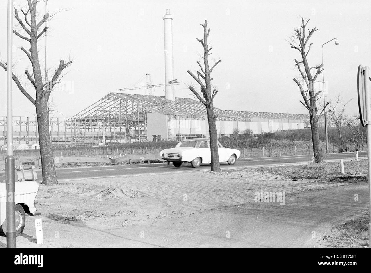 Estensione Hoogovens Wijk a/Zee Hoogovens, Whizgle News, Dutch Desk, Paesi Bassi, 1950 - 2000 il 29-01-1968. L'immagine include questi argomenti. La scena presenta un paesaggio aspro e industriale, dominato da un edificio parzialmente costruito, che rivela un quadro di travi in acciaio. Questa struttura incombe sullo sfondo, con un camino alto che emerge da un lato, suggerendo un sito attivo di industria o produzione. In primo piano si estende una strada, con tratti di ghiaia e larghe sezioni pavimentate su entrambi i lati, che indicano un passaggio da urbano a industriale. Lungo la strada, due alberi Foto Stock
