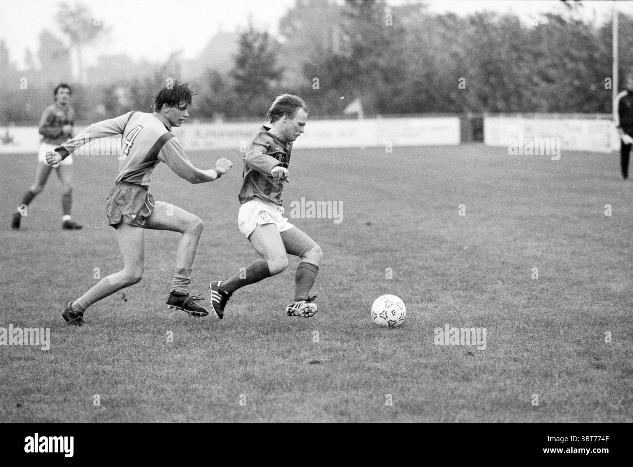 Lisse - Gouda SC Lisse Football, Whizgle News, Dutch Desk, Paesi Bassi, 1950 - 2000 il 17-09-1983. Questi sono gli argomenti nell'immagine. Su un campo sportivo erboso, due giocatori sono impegnati in un intenso momento di una partita di calcio. La scena è ambientata in un ambiente leggermente coperto, con un cielo grigio che indica la possibilità di pioggia. L'erba è lussureggiante e verde, a indicare un campo ben tenuto, punteggiato di macchie di fango del gioco precedente. Il primo giocatore che indossa una maglia arancione brillante è in movimento e lancia abilmente una palla da calcio bianca. Ha dei capelli corti e duri che suggeriscono un senso Foto Stock