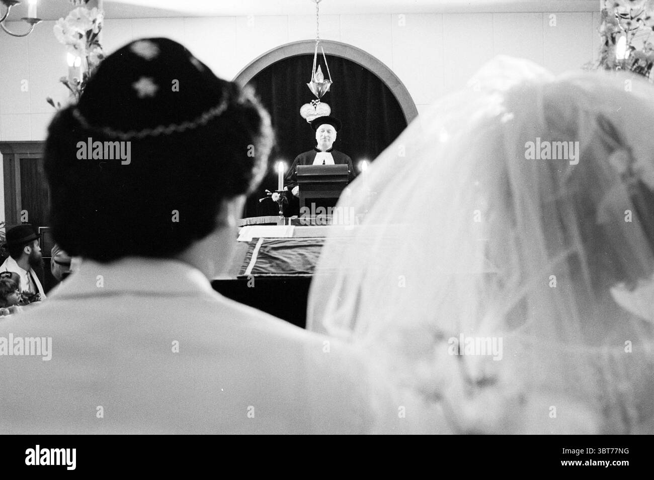 Matrimonio ebraico sinagoga matrimonio, Whizgle News, Dutch Desk, Paesi Bassi, 1950 - 2000 il 21-06-1981. Questi sono gli argomenti nell'immagine. In un ambiente interno molto illuminato, una coppia si trova in prima linea, con le spalle rivolte verso lo spettatore. Lo sposo è rivestito in una tuta bianca, con i caratteristici capelli scuri, ricoperta da un kippah nero adornato da un motivo. Accanto a lui, la sposa è elegantemente vestita con un abito bianco che scorre, il suo velo morbido che le cade sulla schiena e inquadra parzialmente il viso. Sono visibili delicate composizioni floreali, che esaltano delicatamente l'atmosfera romantica. Sullo sfondo, una figura Foto Stock