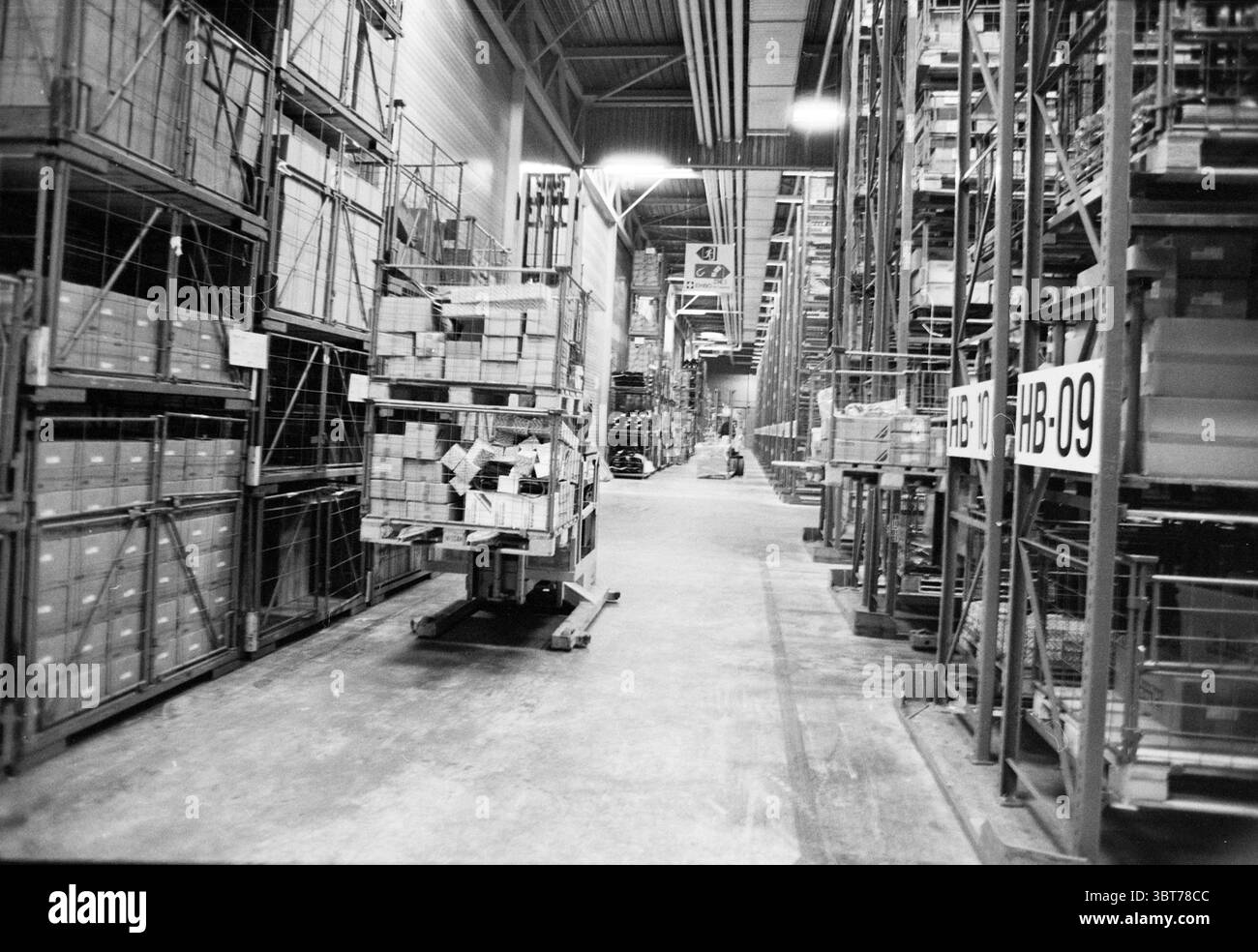 Int Nissan warehouse Amsterdam, Whizgle News, Dutch Desk, Paesi Bassi, 1950 - 2000 on 30-01-1991. Questi sono gli elementi dell'immagine. La scena raffigura un magazzino spazioso pieno di scaffali torreggianti e scaffali, creando un senso di verticalità. La prospettiva attira l'occhio lungo un lungo corridoio, affiancato da scaffali metallici che si estendono verso il soffitto, dove gli apparecchi di illuminazione industriali illuminano l'area con un bagliore fresco e forte. Il pavimento è realizzato in cemento, che appare leggermente usurato e contrassegnato dal movimento delle merci, contribuendo a creare un'atmosfera industriale. Lungo Foto Stock