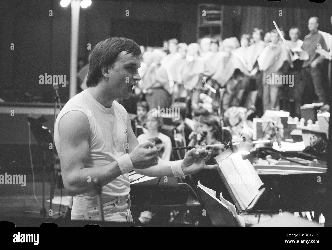 Three sous operetta Music Theatre Opera operetta, Whizgle News, Dutch Desk, Paesi Bassi, 1950 - 2000 il 18-04-1983. L'immagine mostra questi argomenti. La scena presenta un ambiente dinamico pieno di musicisti e cantanti. In primo piano, un direttore d'orchestra si erge con fiducia, gestendo animatamente con le braccia, suggerendo uno stile di leadership entusiasta. Indossa un top senza maniche, enfatizzando il suo fisico, e braccialetti bianchi che aggiungono un tocco sportivo. Dietro di lui, un grande coro è disposto in un semicerchio, le loro teste girano leggermente verso il direttore mentre si impegnano nel perf Foto Stock