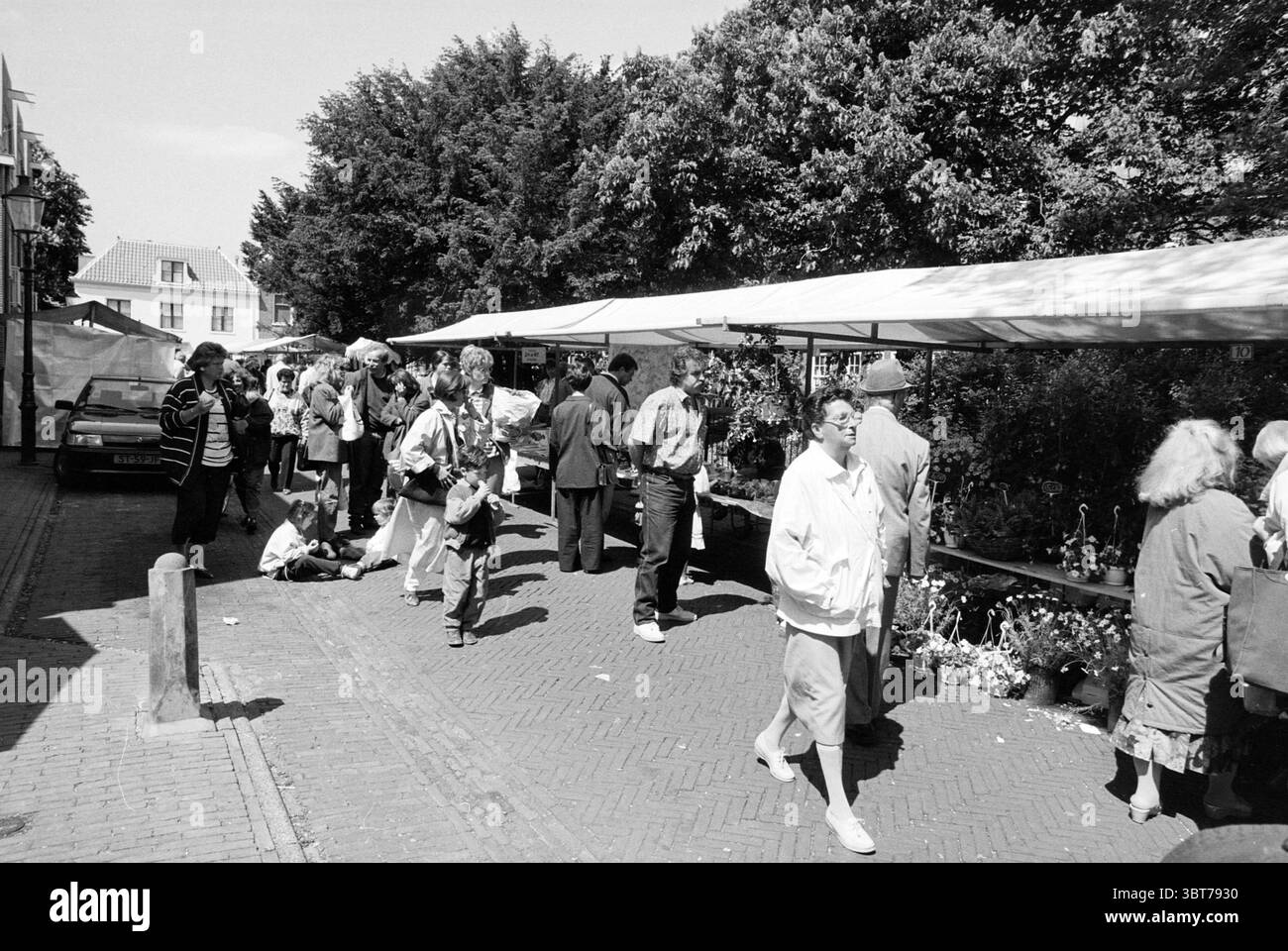 Spring Market Bloemendaal Bloemendaal, Whizgle News, Dutch Desk, Paesi Bassi, 1950 - 2000 il 19-05-1990. L'immagine contiene questi argomenti. La scena raffigura una vivace strada del mercato sotto un cielo luminoso. Una varietà di bancarelle rivestite di baldacchini bianchi creano un senso di organizzazione in un'atmosfera vivace. Le persone sono sparse in tutto lo spazio, impegnate in varie attività, alcune stanno navigando tra le bancarelle mentre altre stanno conversando, creando un'energia vibrante. In primo piano, un bambino siede sul marciapiede di ciottoli, apparentemente intrattenuto dall'ambiente circostante. Nelle vicinanze si trovano anche adulti Foto Stock