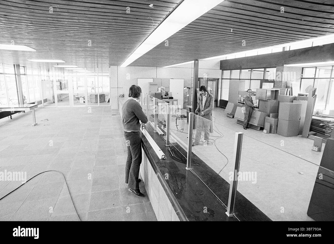 Nuovo edificio HD e ufficio postale, Whizgle News, Dutch Desk, Paesi Bassi, 1950 - 2000. Questi sono gli elementi dell'immagine. La scena raffigura un ampio spazio interno aperto caratterizzato da un design architettonico moderno. Il pavimento e' composto da grandi piastrelle di pietra, che contribuiscono ad un'estetica spaziosa e pulita. Il soffitto presenta una serie di pannelli in legno a doghe, che aggiungono calore all'ambiente altrimenti minimalista. Sul lato sinistro, c'è un bancone evidenziato da una superficie scura ed elegante, che suggerisce un'area di ricevimento o di servizio. Questo contatore è separato dal resto del ro Foto Stock