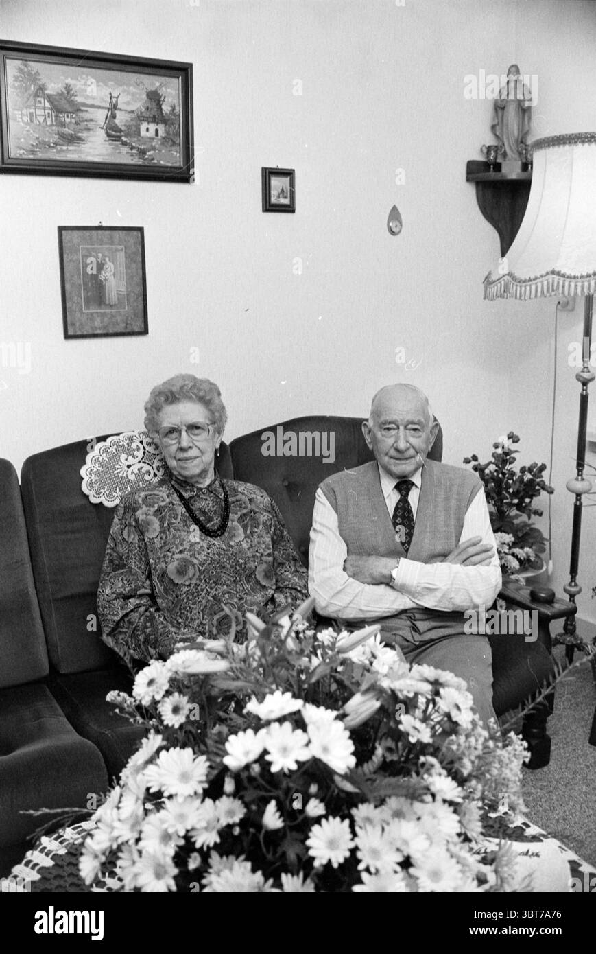60 anni famiglia Willemse, Whizgle News, Dutch Desk, Paesi Bassi, 1950 - 2000 il 07-11-1991. Questi sono gli elementi dell'immagine. In un interno accogliente, una coppia di anziani siede fianco a fianco su un divano imbottito scuro, le loro espressioni trasmettono un senso di compagnia tranquilla. La donna di sinistra ha un atteggiamento premuroso, con i suoi capelli grigi e ricciolati che incorniciano il viso. Indossa un maglione a motivi geometrici che presenta un mix di tonalità terrose: Marroni, verdi e accenni di arancio. Oltre a lei, l'uomo ha un'espressione più neutra, vestita con un gilet di colore chiaro su una camicia con colletto. Il suo calvo Foto Stock