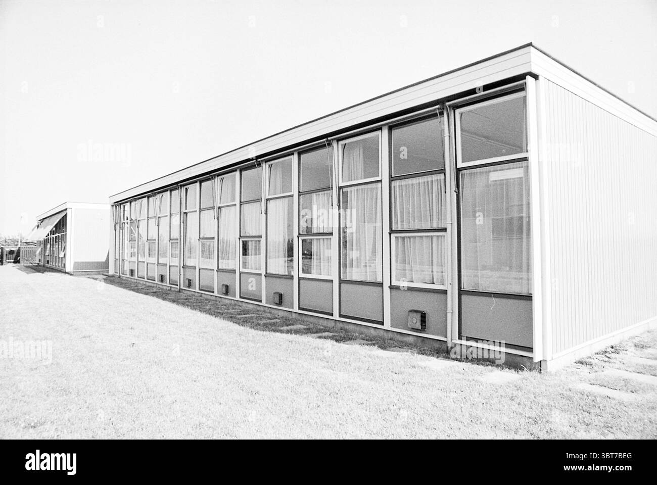 Edifici temporanei edifici di emergenza locali, Whizgle News, Dutch Desk, Paesi Bassi, 1950 - 2000 nel 1975. Questi argomenti vengono visualizzati nell'immagine. La scena presenta un edificio moderno caratterizzato dalla sua architettura elegante e lineare. La struttura presenta una serie di grandi pannelli di vetro allineati simmetricamente su un lato, consentendo alla luce naturale di inondare l'interno, mentre il lato opposto appare più solido con una finitura esterna liscia. La facciata dell'edificio è prevalentemente di colore tenue, probabilmente una combinazione di grigio chiaro e bianco, che contrastano dolcemente con il vibrante verde dell'erba Foto Stock
