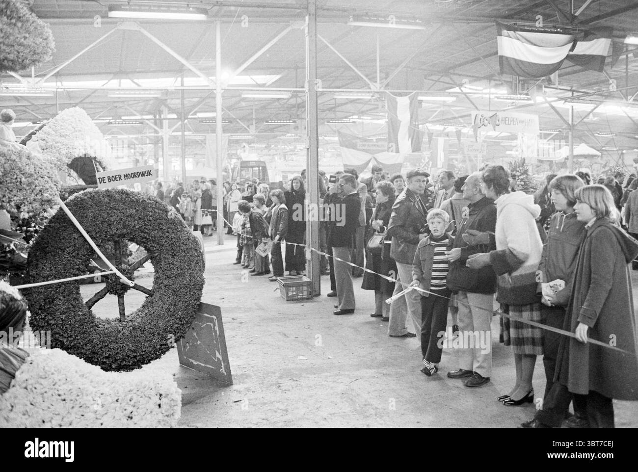 La parata galleggia a Hobaho Lisse Flower Parade mostra di fiori Lisse, Whizgle News, Dutch Desk, Paesi Bassi, 1950 - 2000 il 20-04-1979. Questi sono gli elementi dell'immagine. In un vivace mercato al coperto, si sviluppa una vivace scena. L'atmosfera e' vivace, piena di eccitazione mentre una grande folla si riunisce. La composizione presenta un mix di adulti e bambini, tutti rivolti verso un display centrale. In primo piano, spicca un'ampia disposizione floreale, realizzata a forma di una tradizionale ruota del carrello. I fiori, che potrebbero essere di colore brillante, mostrano una varietà Foto Stock