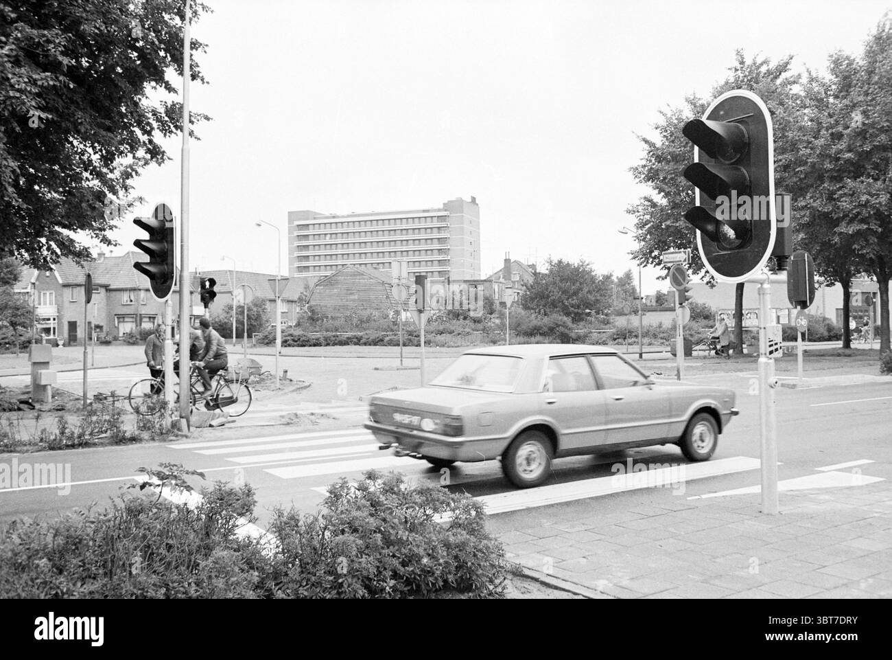 Incrocio con attraversamento pedonale e semafori, Whizgle News, Dutch Desk, Paesi Bassi, 1950 - 2000 nel 1978. L'immagine mostra questi argomenti. La scena presenta un ambiente urbano discreto, caratterizzato da cieli sovrastati che proiettano una luce soffusa e diffusa sull'area. In primo piano, un'auto grigia si muove lungo la strada asfaltata, con la sua forma essenziale che enfatizza il senso di movimento. Il colore del veicolo si fonde armoniosamente con le tonalità silenziose dell'ambiente. A sinistra, due persone in bicicletta sono posizionate vicino a un segnale stradale, alto e imponente con li verde brillante Foto Stock