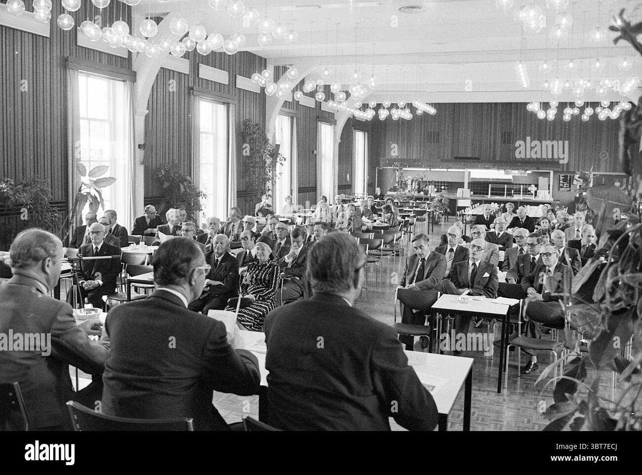 200 anni di esistenza di Ned.Mij Handel en Nijverh. Ricevimenti reali e visite reali, Whizgle News, Dutch Desk, Paesi Bassi, 1950 - 2000 il 09-06-1977. Questi sono gli elementi dell'immagine. La scena presenta uno spazioso ambiente interno con pavimento in legno lucido e grandi finestre che lasciano inondare l'area con luce naturale. Le pareti sono adornate con caldi pannelli marroni, contribuendo a creare un'atmosfera intima. Sospesi dal soffitto sono molteplici lampade rotonde e luminose che emettono un'illuminazione delicata, migliorando l'atmosfera invitante dello spazio. In primo piano, un gruppo di dresse formalmente Foto Stock