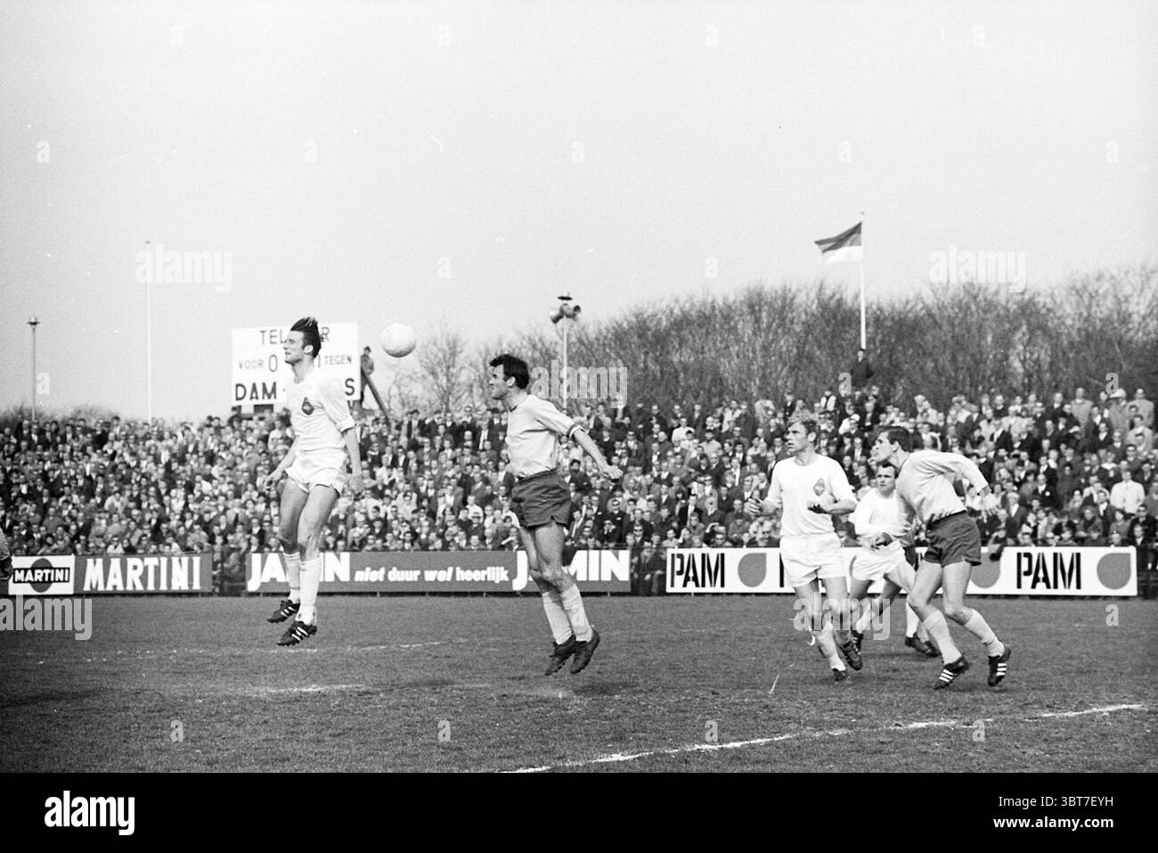 Calcio maschile Telstar., Whizgle News, Dutch Desk, Paesi Bassi, 1950 - 2000. L'immagine include questi argomenti. Sulla scena, due squadre sono impegnate in una vivace partita di calcio su un campo erboso, circondato da una folla di spettatori. I giocatori mostrano una varietà di espressioni mirate mentre competono per la palla, che è trasportata in aria e sembra essere diretta verso uno dei giocatori che saltano per intercettarla. Il giocatore in bianco, con la maglia numero 9, è in posizione di mezz'aria, braccia allungate e occhi bloccati sulla palla, mostrando un momento di atleticità. I suoi compagni di squadra e gli avversari sono in vari punti Foto Stock