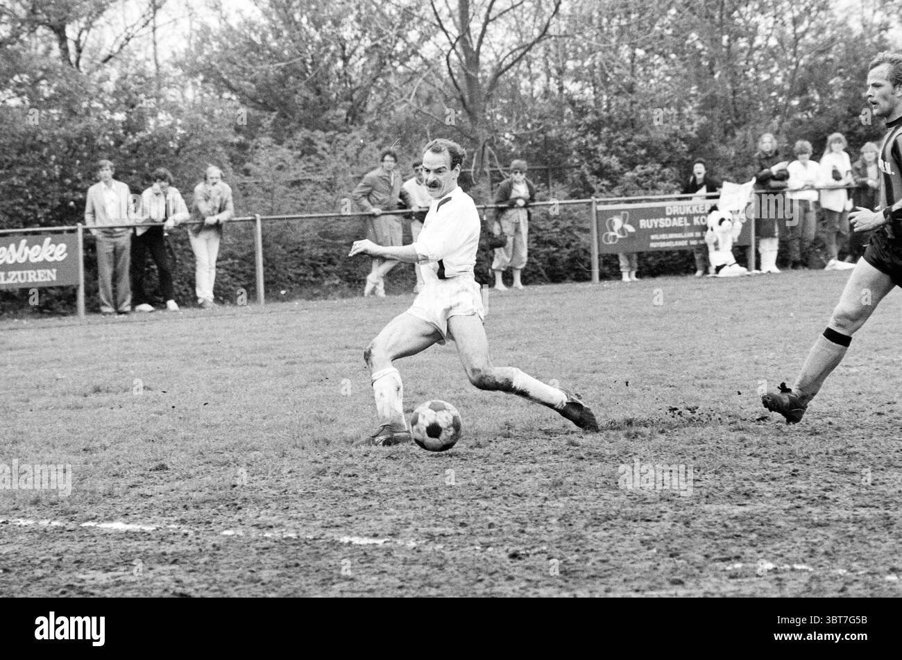 Partita di calcio tra Harsveld Velsen e Buys B.v., Whizgle News, Dutch Desk, Paesi Bassi, 1950 - 2000 nel 1983. L'immagine include questi argomenti. Un campo da calcio erboso si estende lungo tutta la scena, con la sua superficie segnata da macchie fangose provenienti da partite recenti. In primo piano, un giocatore con una maglia bianca e pantaloncini corti è in azione, inclinandosi in avanti con un'espressione decisa, mentre manovra abilmente la palla con il piede. Il suo corpo è leggermente angolato lateralmente, mostrando l'intensità del gioco. A destra, un secondo giocatore, rivestito di pantaloncini scuri e una maglia a strisce, si prepara a fare un poss Foto Stock