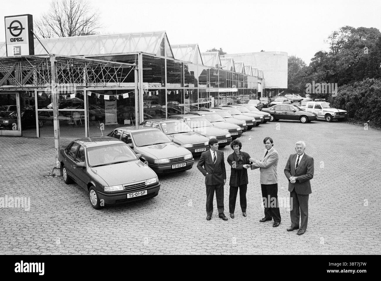 Azienda automobilistica, Whizgle News, Dutch Desk, Paesi Bassi, 1950 - 2000 il 10-1988. Questi argomenti vengono visualizzati nell'immagine. In un ambiente spazioso all'aperto, una concessionaria di auto presenta una disposizione ordinata dei veicoli. Il primo piano presenta una fila di auto dall'aspetto simile, principalmente in tonalità di grigio e argento, parcheggiate l'una vicino all'altra, suggerendo un'estetica della metà degli anni '90. Di fronte a queste auto ci sono quattro persone, tutte vestite con abbigliamento formale, che suggeriscono un'atmosfera professionale. Il loro linguaggio del corpo è aperto e coinvolgente; una persona fa gesti come se facesse un punto, mentre un'altra ascolta attentamente. L'altro Foto Stock