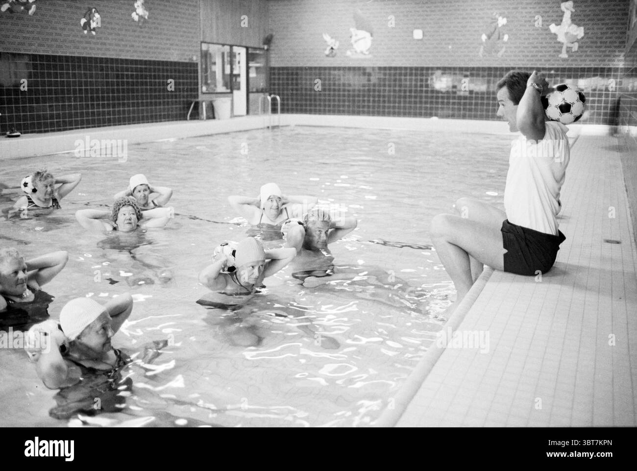Piscina Aquagym, Whizgle News, Dutch Desk, Paesi Bassi, 1950 - 2000 al 05-1989. L'immagine mostra questi argomenti. La scena è ambientata in un'area piscina, caratterizzata dalle sue pareti piastrellate e dall'acqua limpida che riflette l'illuminazione sopraelevata. L'atmosfera è energica e concentrata, illuminata da luci artificiali luminose che creano un'atmosfera calda e vivace. In primo piano, un gruppo di diversi individui, principalmente donne, sono impegnati in una routine di esercizi acquatici. Indossano copricapo corrispondenti e sembrano essere in movimento sincronizzato, sollevando oggetti sopra la testa con la concentrazione Foto Stock
