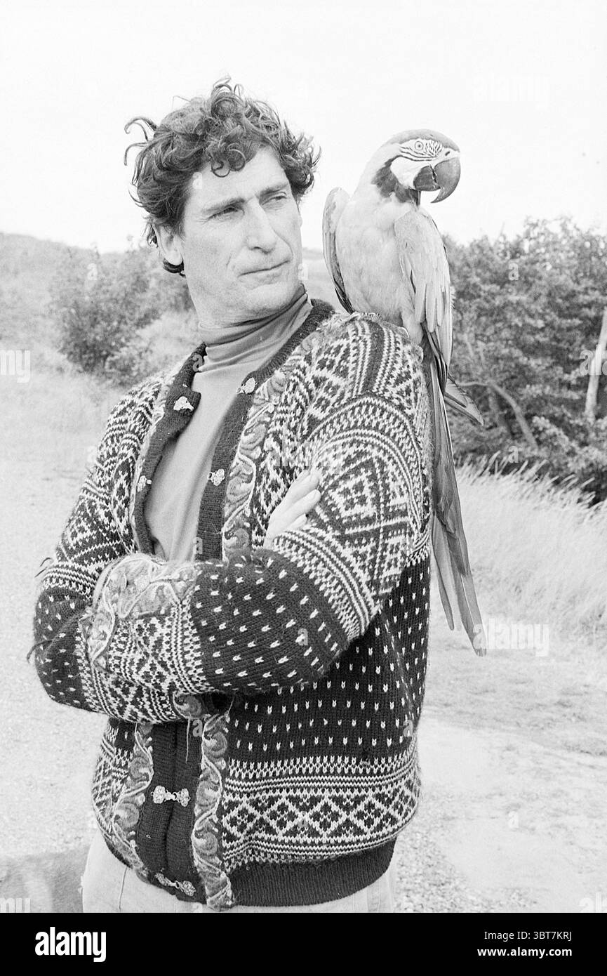Brandwijk con pappagalli, Whizgle News, Dutch Desk, Paesi Bassi, 1950 - 2000 il 14-09-1977. Questi sono gli argomenti nell'immagine. La scena cattura un uomo in piedi all'aperto con una posa sicura e le braccia incrociate sul petto. La sua espressione è premurosa e leggermente seria, suggerendo contemplazione. Ha i capelli ricci che aggiungono un tocco di informalità al suo comportamento. Avvolto intorno a lui c'è un maglione con disegni intricati in tonalità scure, in contrasto con il paesaggio naturale e più chiaro che si nasconde dietro di lui. Appollaiato sulla sua spalla c'è un pappagallo colorato, che attira l'attenzione con il suo Foto Stock