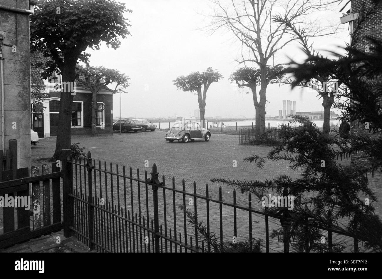 Parcheggio a Oud Velsen con vista sul Canale del Mare del Nord e sulle torri del Velsertunnel, Whizgle News, Dutch Desk, Paesi Bassi, 1950 - 2000. Questi sono gli elementi dell'immagine. La scena presenta un ambiente tranquillo e urbano caratterizzato da un'estetica vintage. In primo piano, una recinzione in ferro battuto delinea una piazza acciottolata, i suoi elementi scuri contrastano con le sfumature più chiare delle strutture circostanti. Un'auto piccola e compatta occupa una posizione centrale nella piazza, con la sua forma arrotondata e le curve morbide che si distinguono contro le linee rigide degli edifici. Fiancheggiando il quadrato, Foto Stock