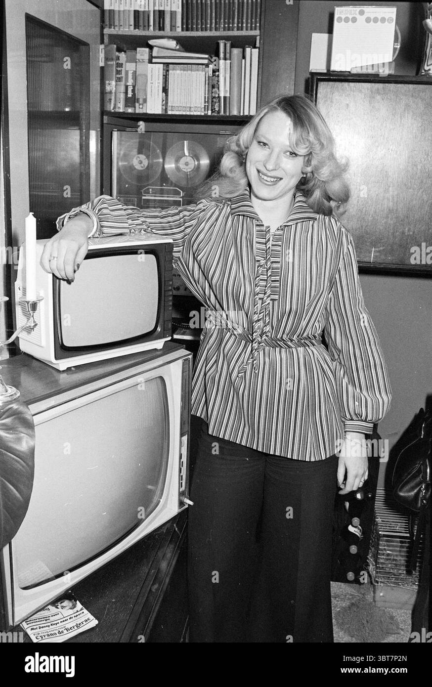 Consegna della TV alla famiglia Bastianen (in connessione con il concorso Belinda) consegna e trasferimenti, Whizgle News, Dutch Desk, Paesi Bassi, 1950 - 2000 il 13-04-1977. Questi argomenti vengono visualizzati nell'immagine. La scena presenta una donna in piedi con sicurezza accanto a due televisori d'epoca, entrambi con un'aura nostalgica. Indossa una blusa a righe con maniche larghe che sfoggia un vivace motivo di colori alternati, prevalentemente con tonalità audaci. La sua acconciatura cade in onde morbide, contribuendo a creare un'atmosfera retrò. Lo sfondo rivela un ambiente accogliente e domestico, Foto Stock Consegna della TV alla famiglia Bastianen (in connessione con il concorso Belinda) consegna e trasferimenti, Whizgle News, Dutch Desk, Paesi Bassi, 1950 - 2000 il 13-04-1977. Questi argomenti vengono visualizzati nell'immagine. La scena presenta una donna in piedi con sicurezza accanto a due televisori d'epoca, entrambi con un'aura nostalgica. Indossa una blusa a righe con maniche larghe che sfoggia un vivace motivo di colori alternati, prevalentemente con tonalità audaci. La sua acconciatura cade in onde morbide, contribuendo a creare un'atmosfera retrò. Lo sfondo rivela un ambiente accogliente e domestico, Foto Stock