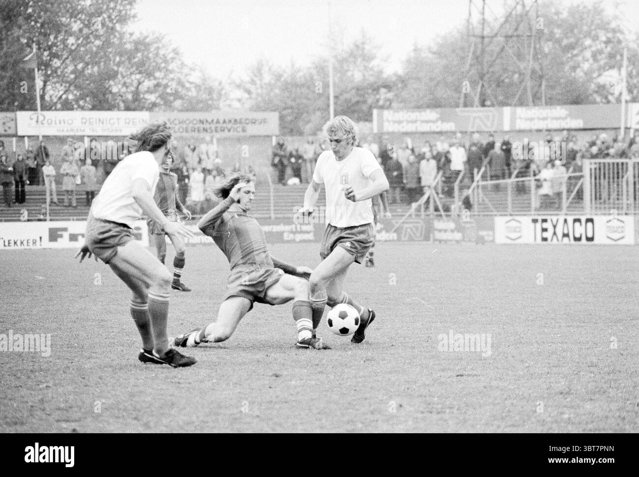 Haarlem - FC Amsterdam Football Haarlem, Whizgle News, Dutch Desk, Paesi Bassi, 1950 - 2000 il 05-05-1974. Questi argomenti vengono visualizzati nell'immagine. In un ambiente all'aperto, la scena cattura un momento energetico di una partita di calcio. Due giocatori di squadre avversarie sono presenti in una posizione centrale, impegnati in un gioco dinamico. Un giocatore che indossa una maglia blu scivola abilmente per intercettare la palla, mostrando agilità e determinazione. L'altro giocatore, con una maglia bianca, è pronto a colpire o manovrare l'avversario che scorre, indicando uno spirito competitivo. Intorno a loro, l'erba f Foto Stock