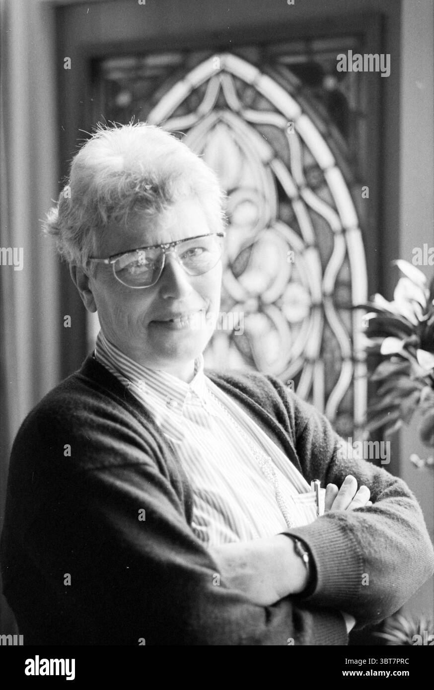 Signora Zuurbier Driehuis Driehuis, Whizgle News, Dutch Desk, Paesi Bassi, 1950 - 2000 al 28-03-1994. L'immagine mostra questi argomenti. In uno spazio interno ben illuminato, un uomo sta in piedi casualmente con le braccia incrociate, trasudando una sicurezza rilassata. Ha i capelli di colore chiaro, leggermente rinforzati, e indossa grandi occhiali rotondi che gli conferiscono un aspetto premuroso. Il suo maglione è scuro, in contrasto con una camicia di colore chiaro sotto che presenta righe orizzontali. Dietro di lui, una finissima finestra colorata filtra la luce soffusa e diffusa, proiettando motivi colorati sulla superficie circostante. T Foto Stock