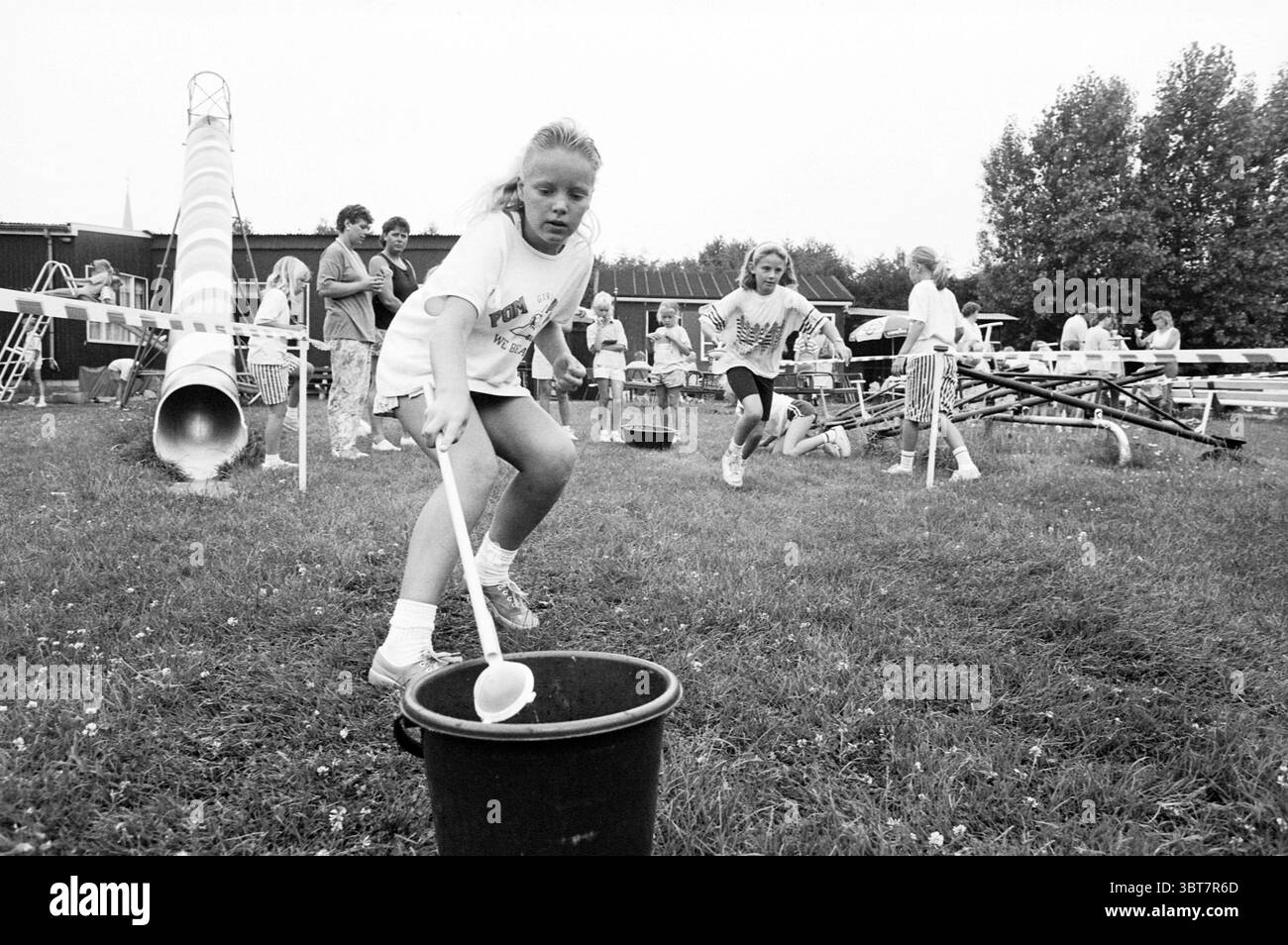 Playground Kindervreugd Hillegom giochi e giocattoli Hillegom, Whizgle News, Dutch Desk, Paesi Bassi, 1950 - 2000 il 08-07-1989. Questi argomenti vengono visualizzati nell'immagine. Una scena vivace si svolge in un ambiente erboso all'aperto, probabilmente parte di un evento della comunità o di una giornata sul campo. Diversi bambini sono impegnati in varie attività, mostrando un senso di competizione e gioia. In primo piano, due ragazze corrono verso un secchio nero. Una ragazza, con lunghi capelli biondi legati, stringe un cucchiaio bianco, pronto a raccogliere qualcosa dal secchio, la sua espressione è concentrata e determinata. Indossa un shi di colore chiaro Foto Stock Playground Kindervreugd Hillegom giochi e giocattoli Hillegom, Whizgle News, Dutch Desk, Paesi Bassi, 1950 - 2000 il 08-07-1989. Questi argomenti vengono visualizzati nell'immagine. Una scena vivace si svolge in un ambiente erboso all'aperto, probabilmente parte di un evento della comunità o di una giornata sul campo. Diversi bambini sono impegnati in varie attività, mostrando un senso di competizione e gioia. In primo piano, due ragazze corrono verso un secchio nero. Una ragazza, con lunghi capelli biondi legati, stringe un cucchiaio bianco, pronto a raccogliere qualcosa dal secchio, la sua espressione è concentrata e determinata. Indossa un shi di colore chiaro Foto Stock